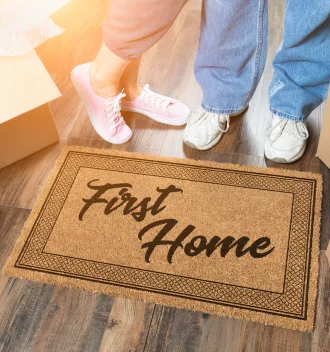 Happy couple standing by a doormat reading "first home," celebrating their first time buying a house